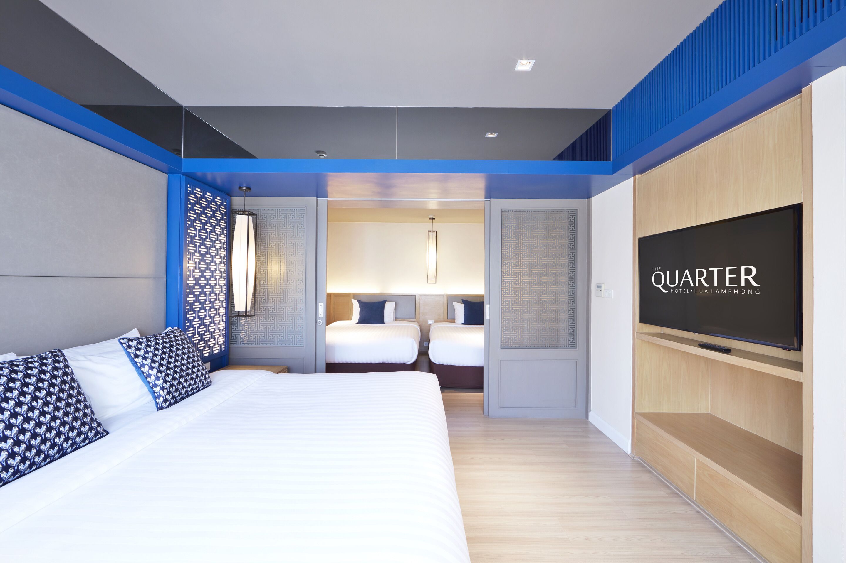A modern hotel room with a large bed, two smaller beds, a TV, and a decorative wall panel.