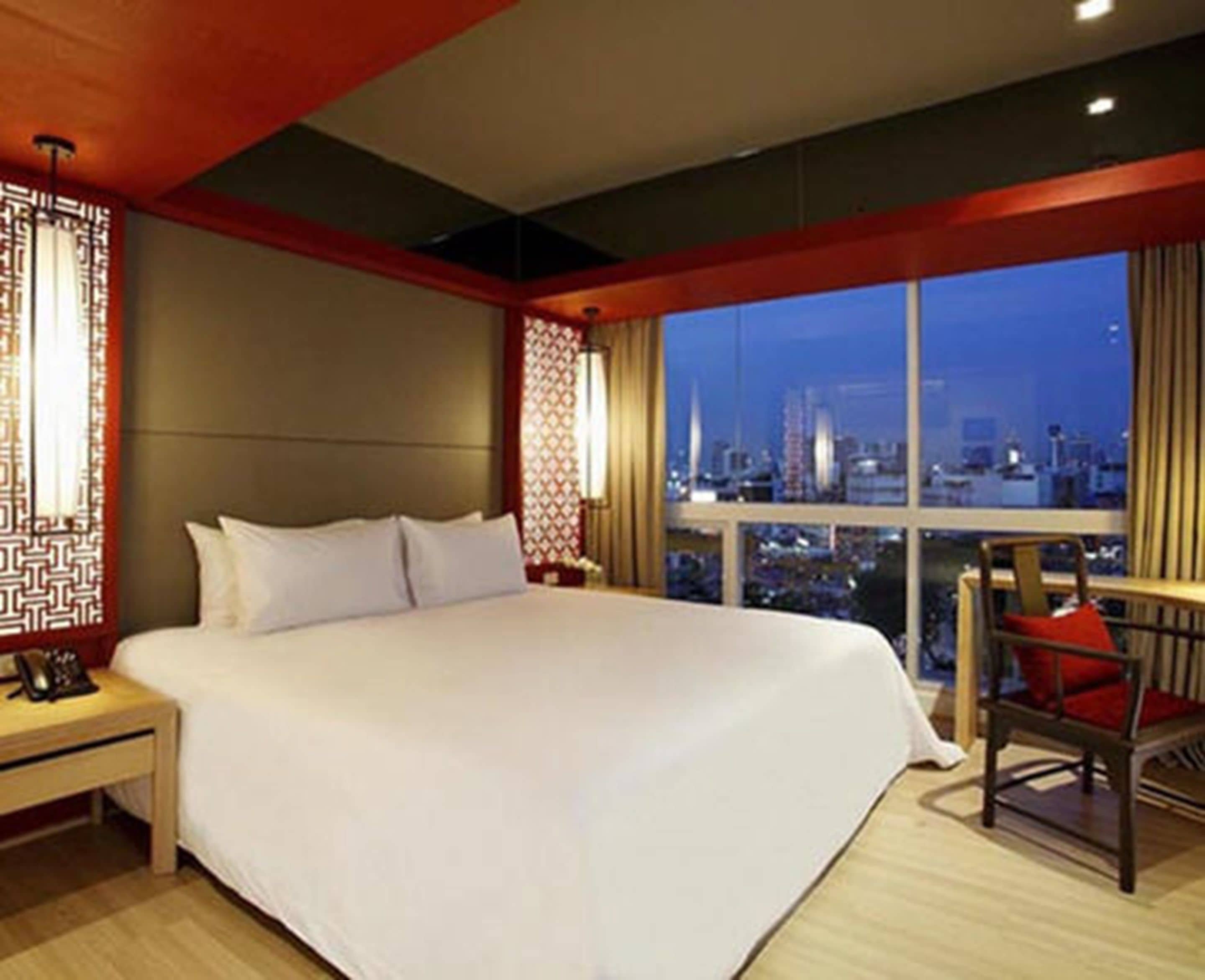 A hotel room with a large bed, a nightstand, a chair, and a view of the cityscape through the window.