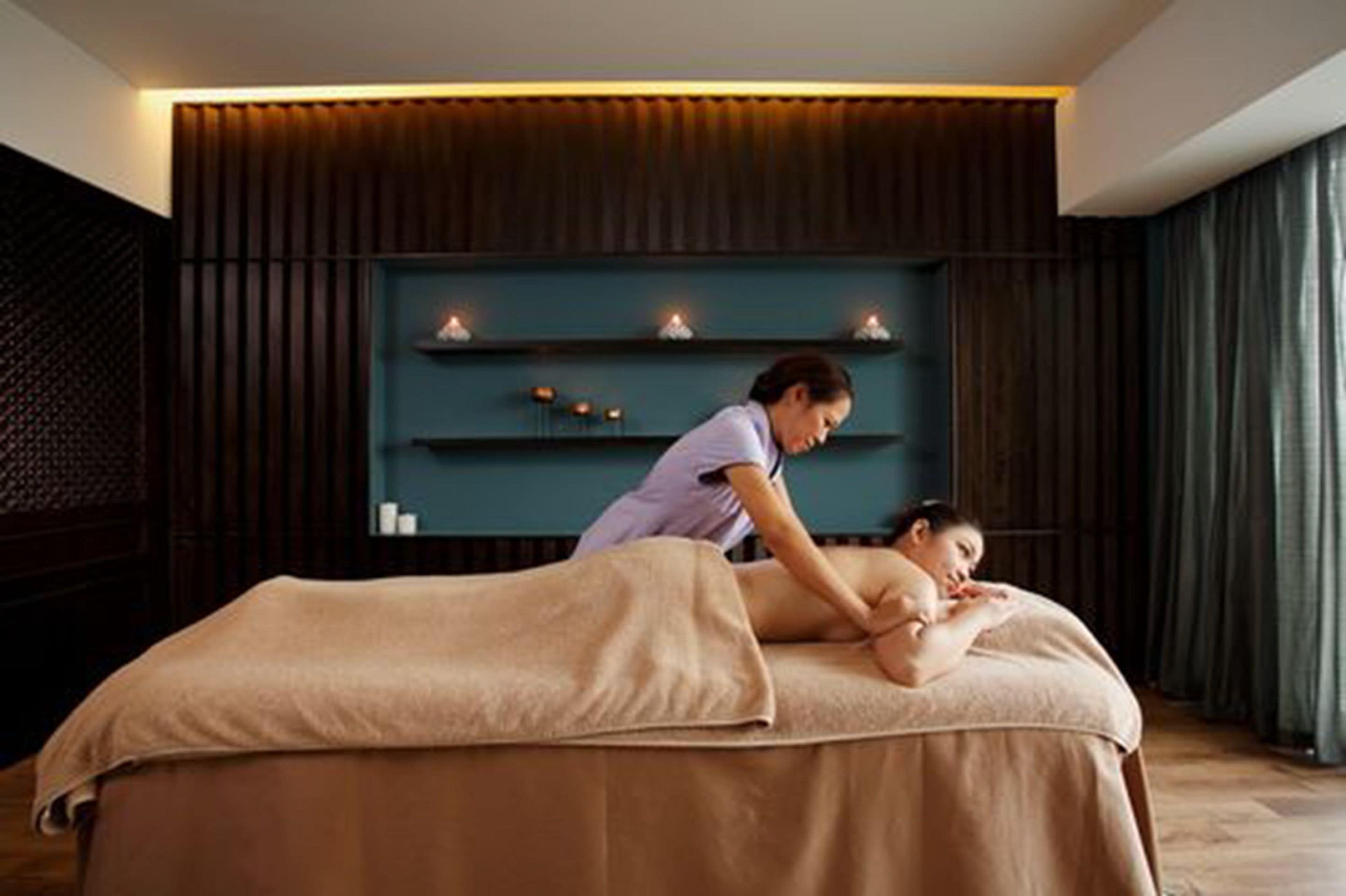 A massage therapy room with a massage table, a person providing a massage, and a shelf with candles in the background.