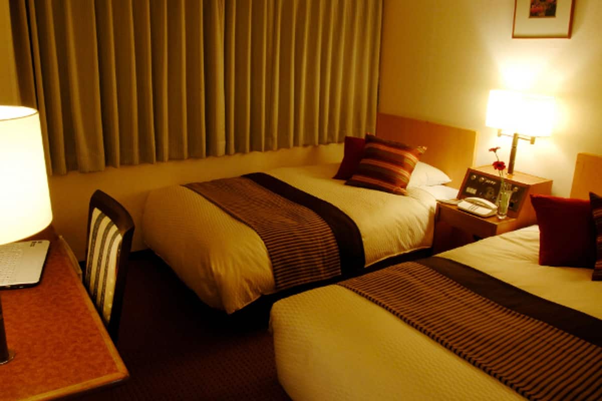 A hotel room with two beds, a desk, a chair, a lamp, and a nightstand with a phone and a vase of flowers.