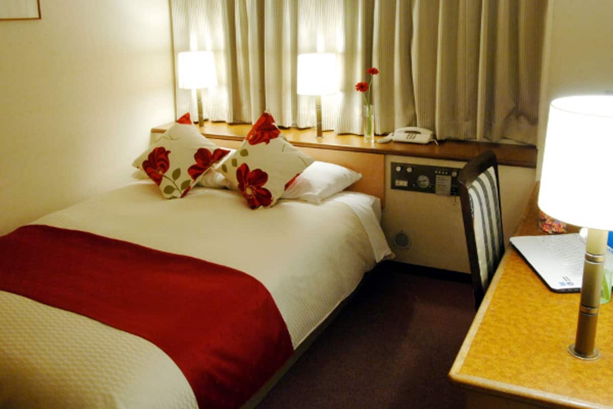 A hotel room with a bed, two bedside lamps, a nightstand with a phone, and a window with curtains.