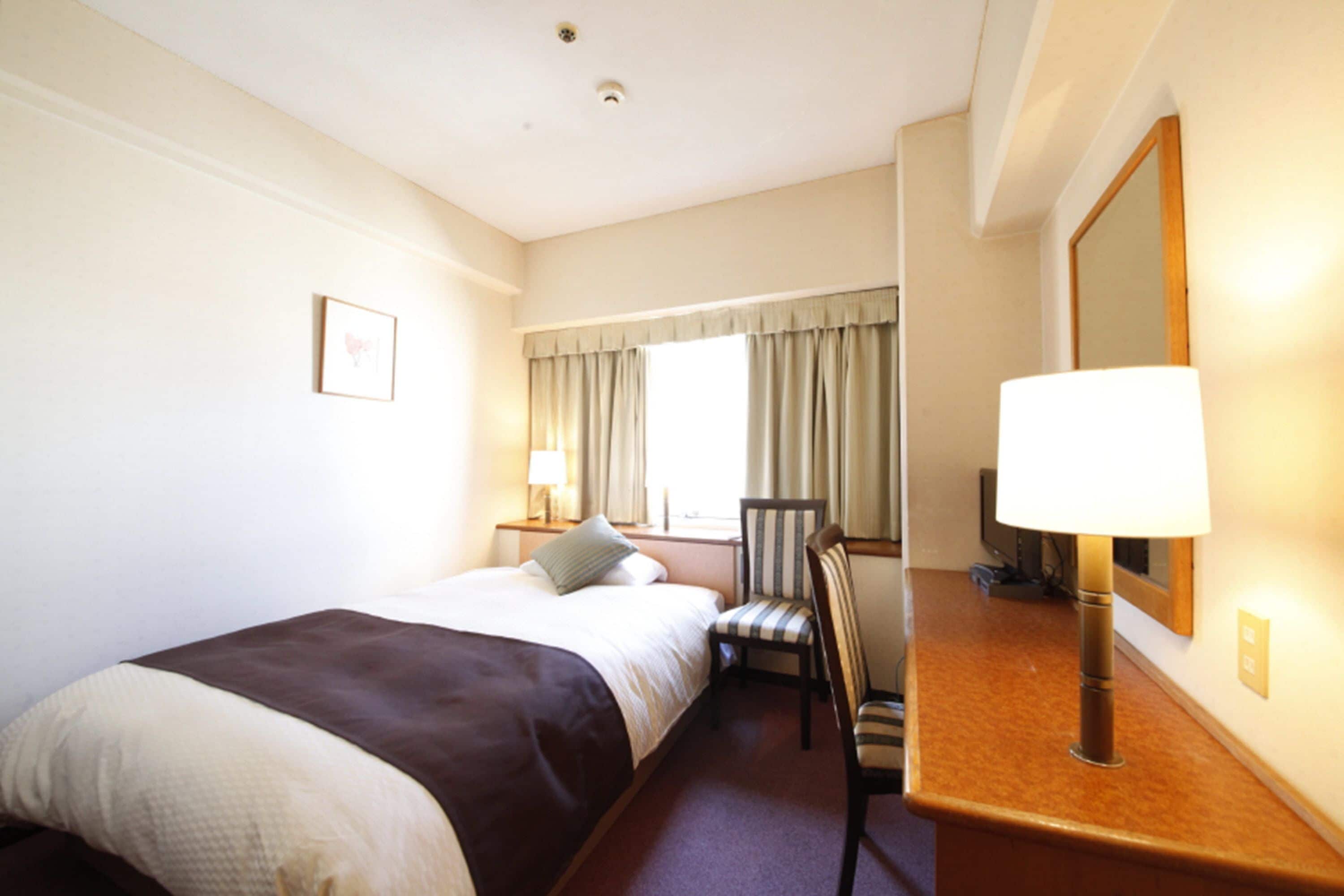 A hotel room with a bed, a chair, a desk with a lamp, a mirror, and a window with curtains.