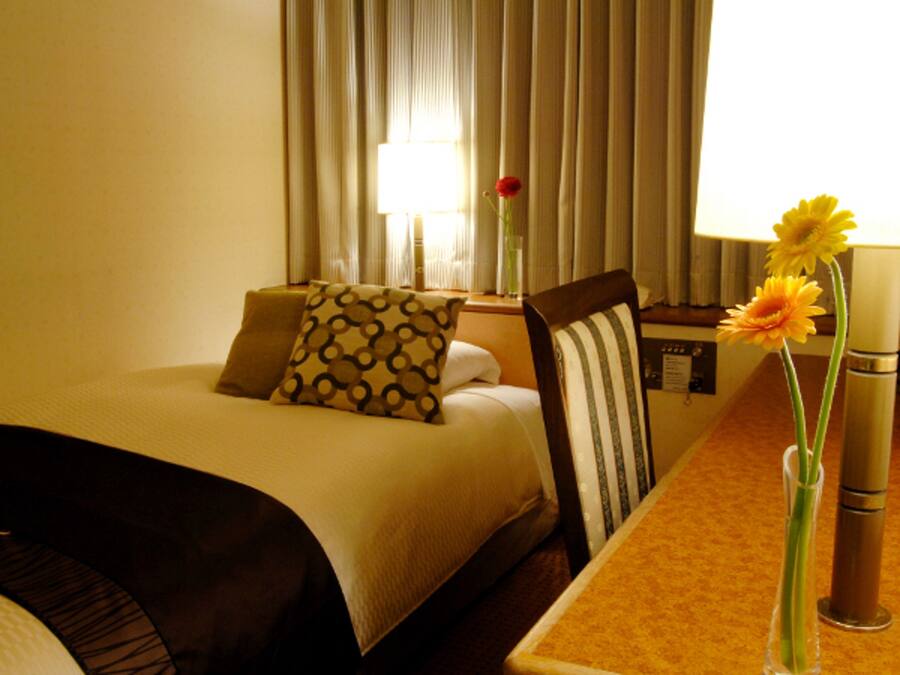 A hotel room with a bed, a chair, a table, and a vase with flowers.