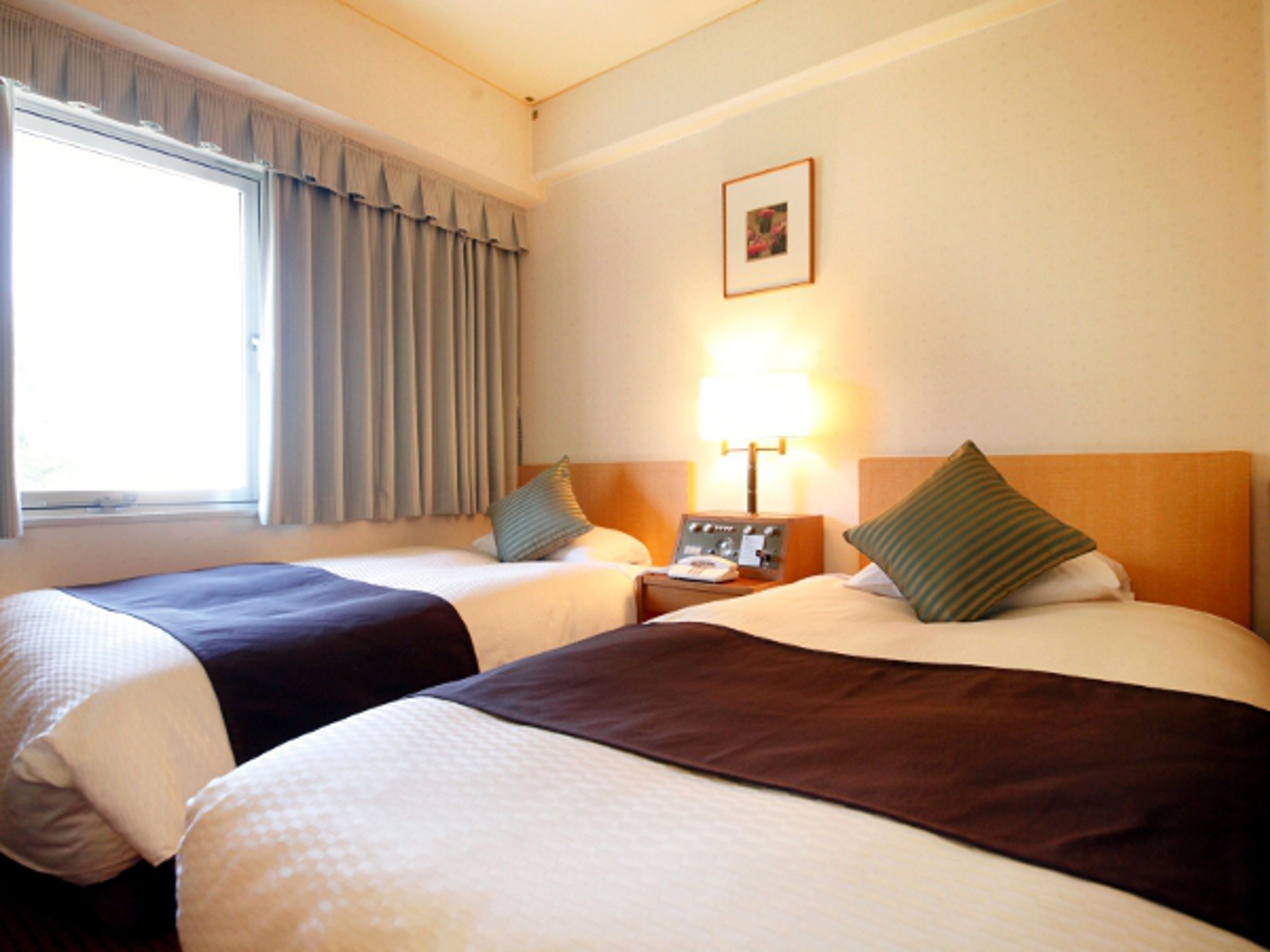 A hotel room with two beds, a window with curtains, a bedside table with a lamp, and a framed picture on the wall.