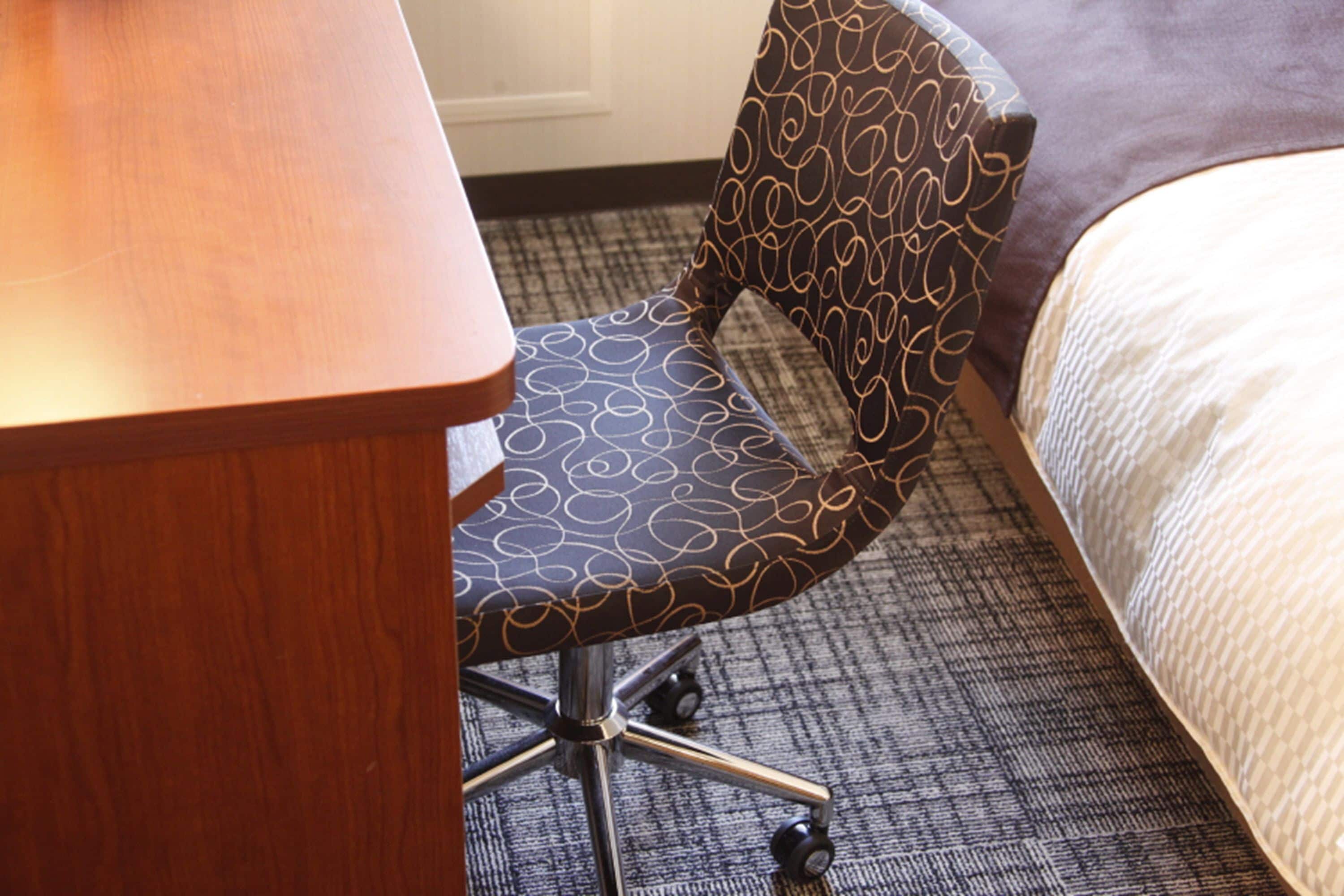 A hotel room with a wooden desk, a patterned armchair, a carpeted floor, and a bed with a white comforter.