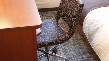 A hotel room with a wooden desk, a patterned armchair, a carpeted floor, and a bed with a white comforter.