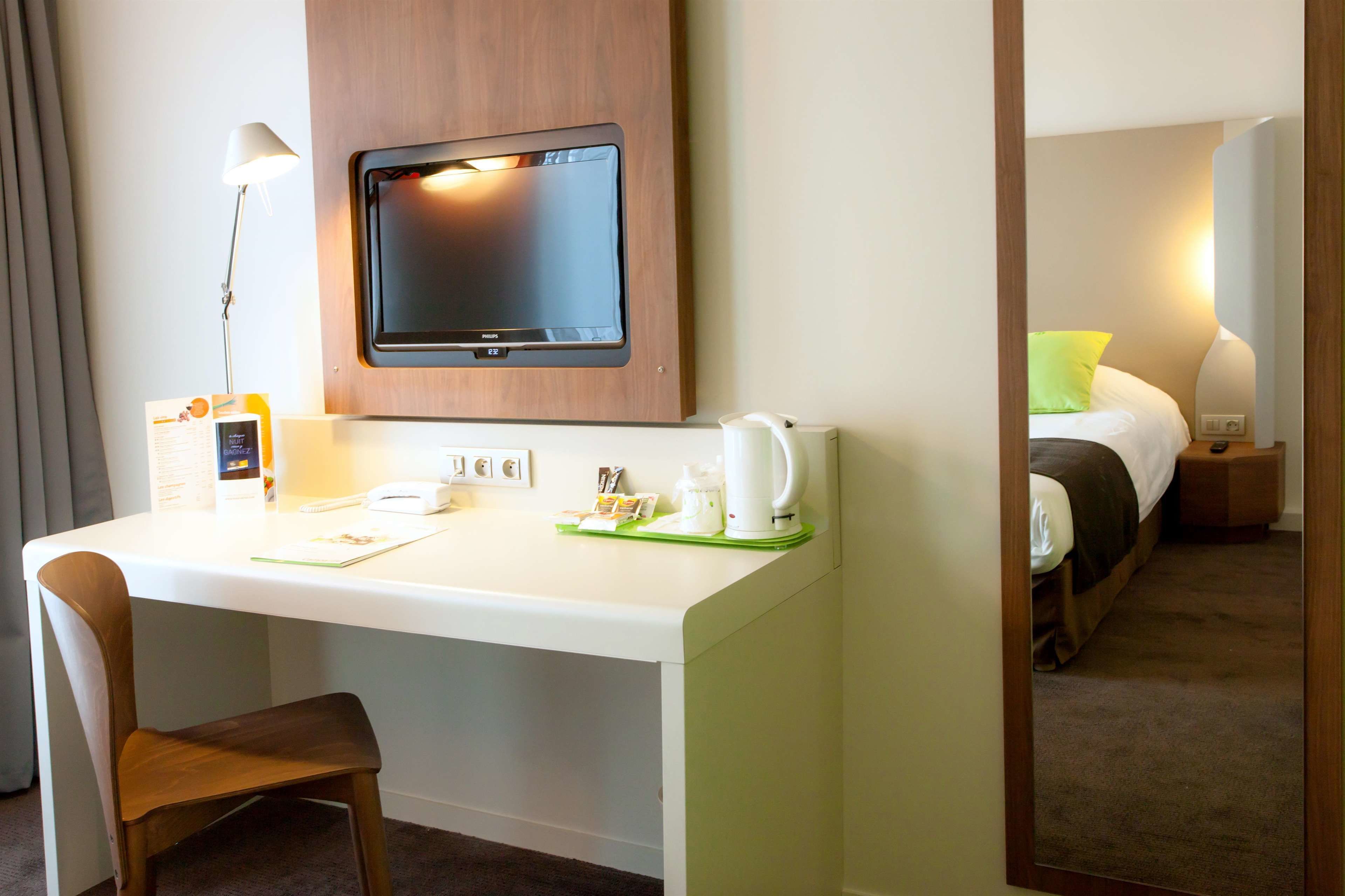 A hotel room with a desk, chair, flat-screen TV, bedside table with a lamp, and a bed with a green pillow.