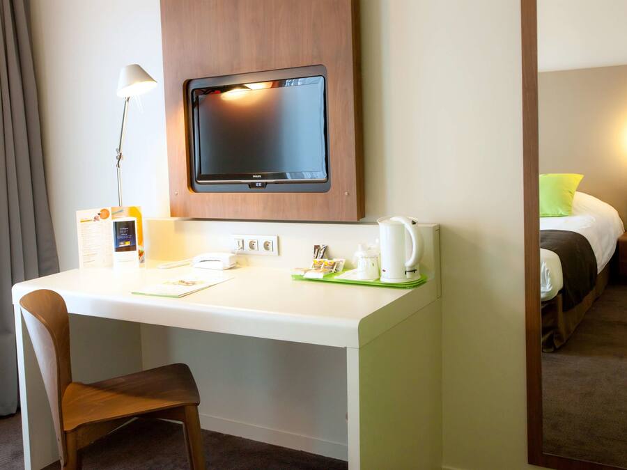 A hotel room with a desk, chair, flat-screen TV, bedside table with a lamp, and a bed with a green pillow.