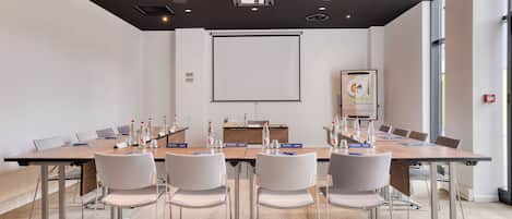 Uma sala de conferência com uma mesa comprida, cadeiras brancas, uma tela de projetor e um quadro branco.