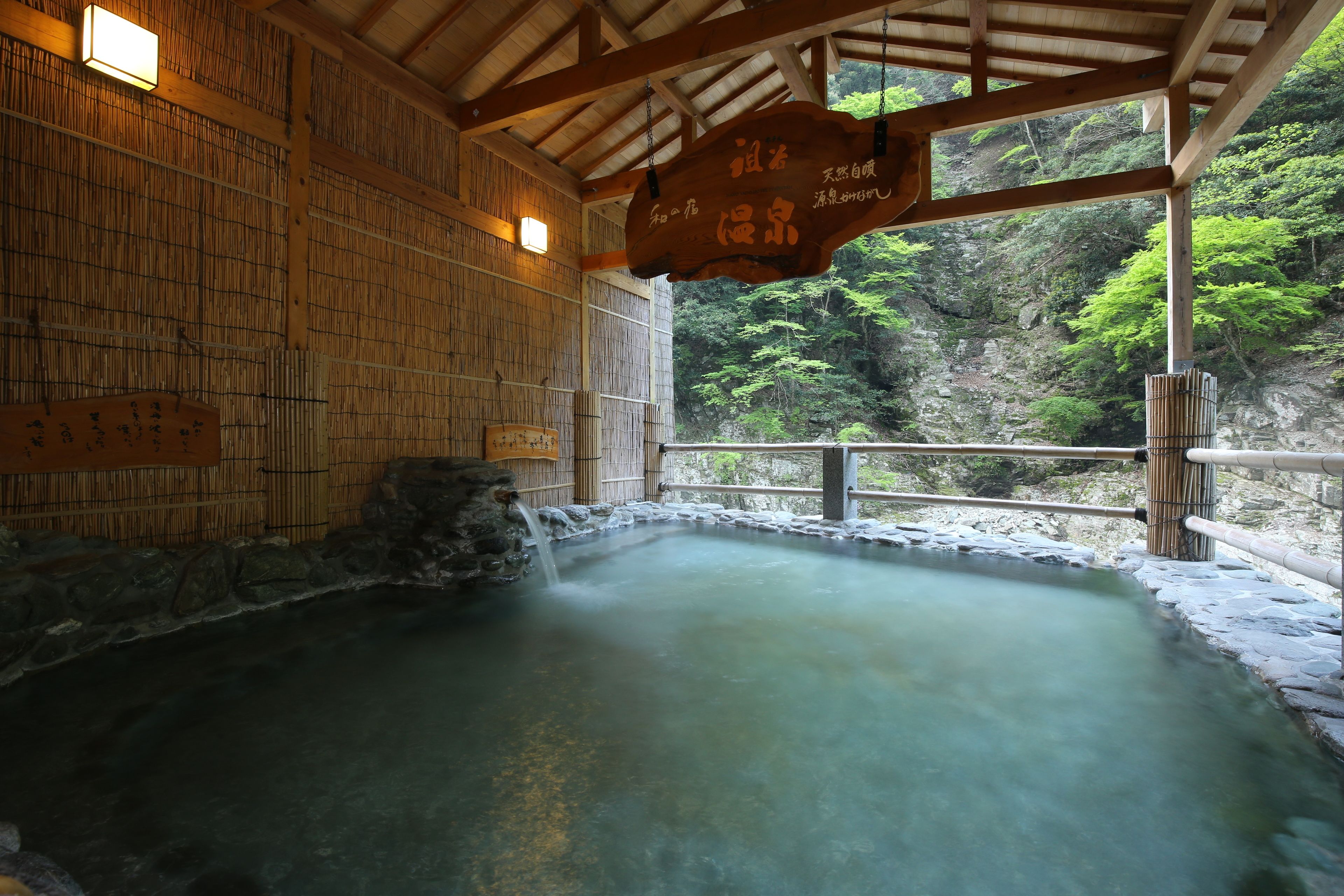 Ein traditionelles japanisches Onsen mit Holzvertäfelung, Steinwänden und Blick auf üppiges Grün außerhalb.