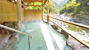 Un onsen japonés tradicional con estructura de madera, escalones de piedra que conducen a una fuente termal cristalina y vistas a una ribera rocosa y montañas boscosas.