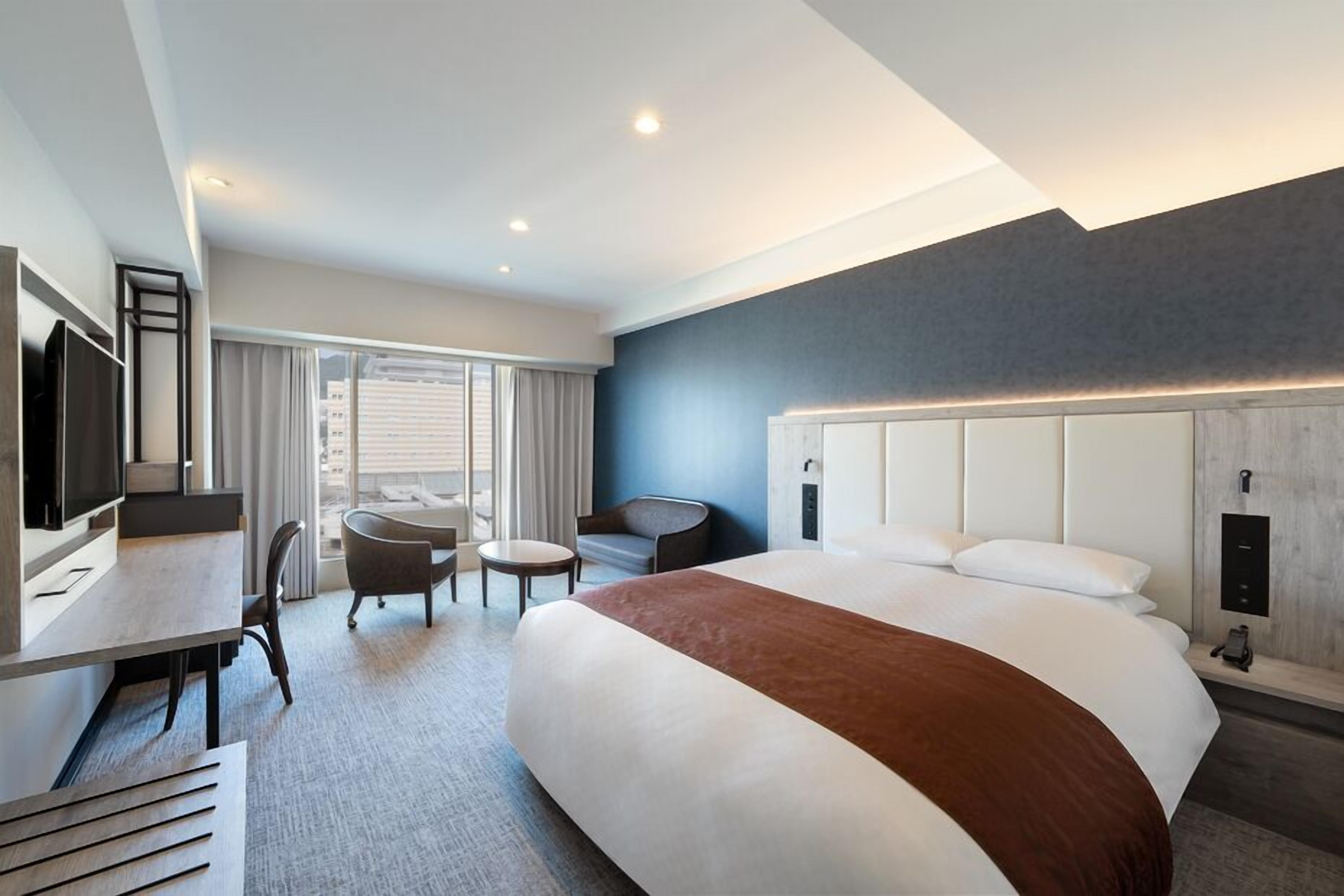 A modern hotel room with a large bed, a desk with a chair, a small round table, and a television.