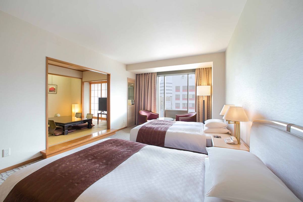 A hotel room with two beds, a desk, a chair, a lamp, and a view of the city.