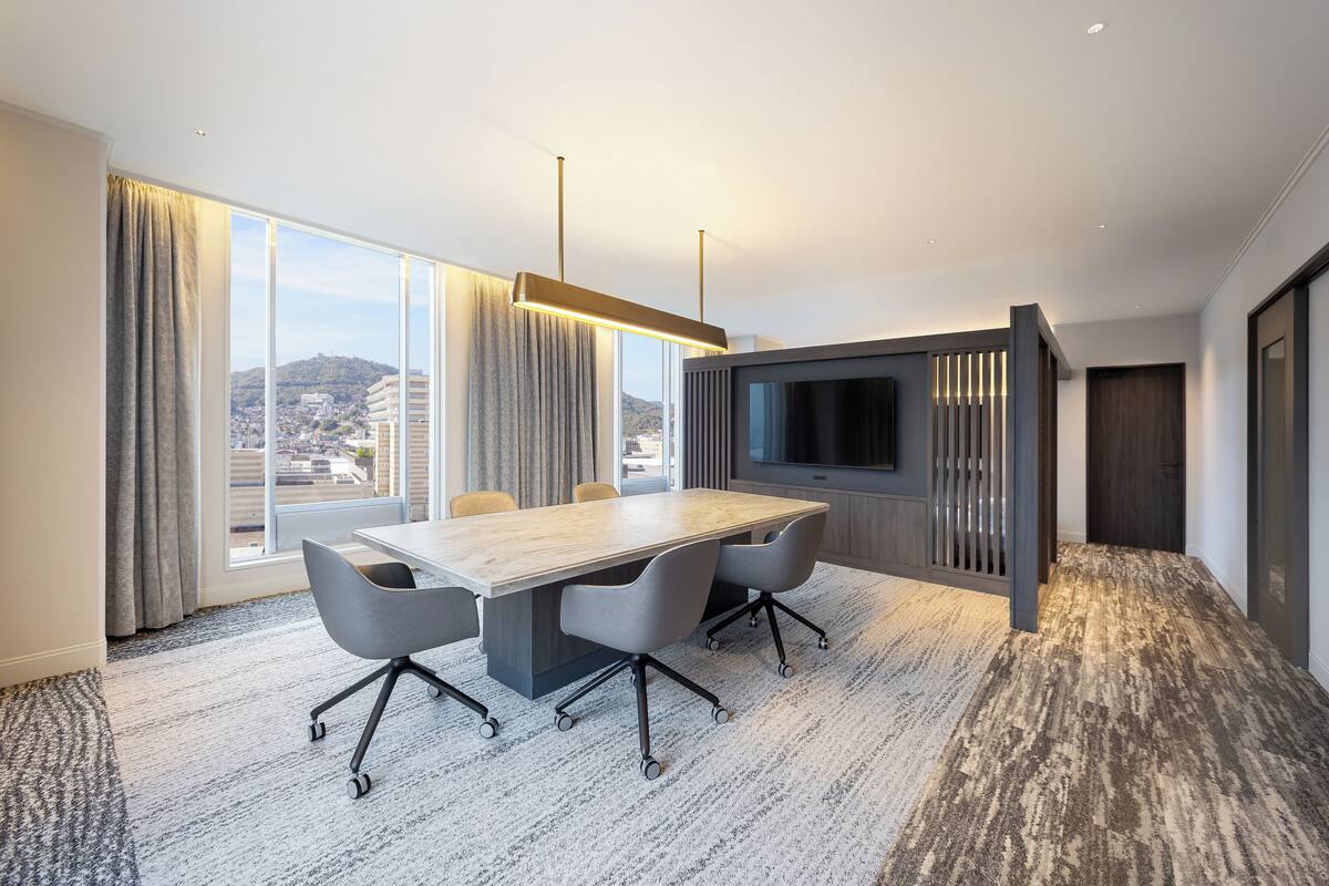 A conference room with a large table, four chairs, a flat-screen TV, and a city view through large windows.