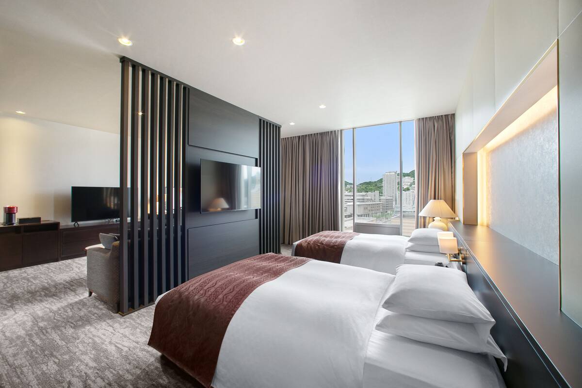 A hotel room with two beds, a TV, a desk, and a view of the city.