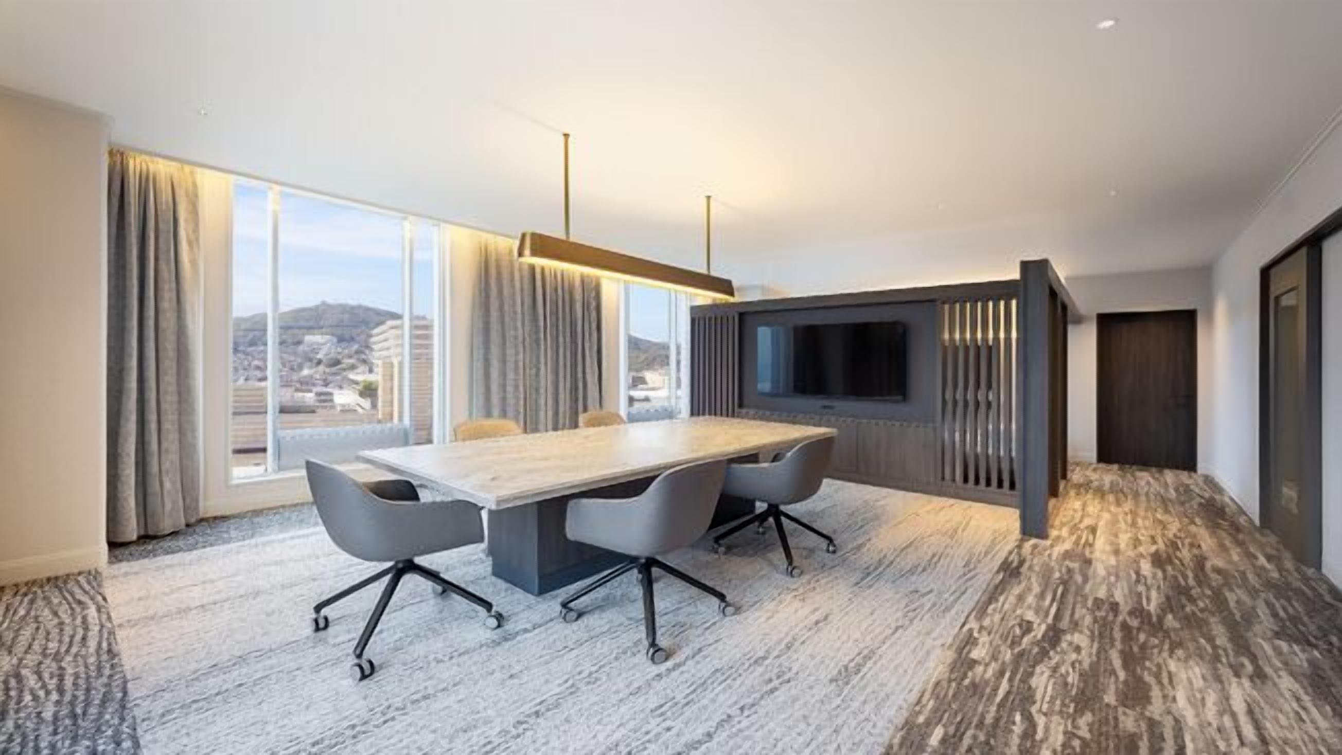 A modern hotel room with a large conference table, a flat-screen TV, and a city view.