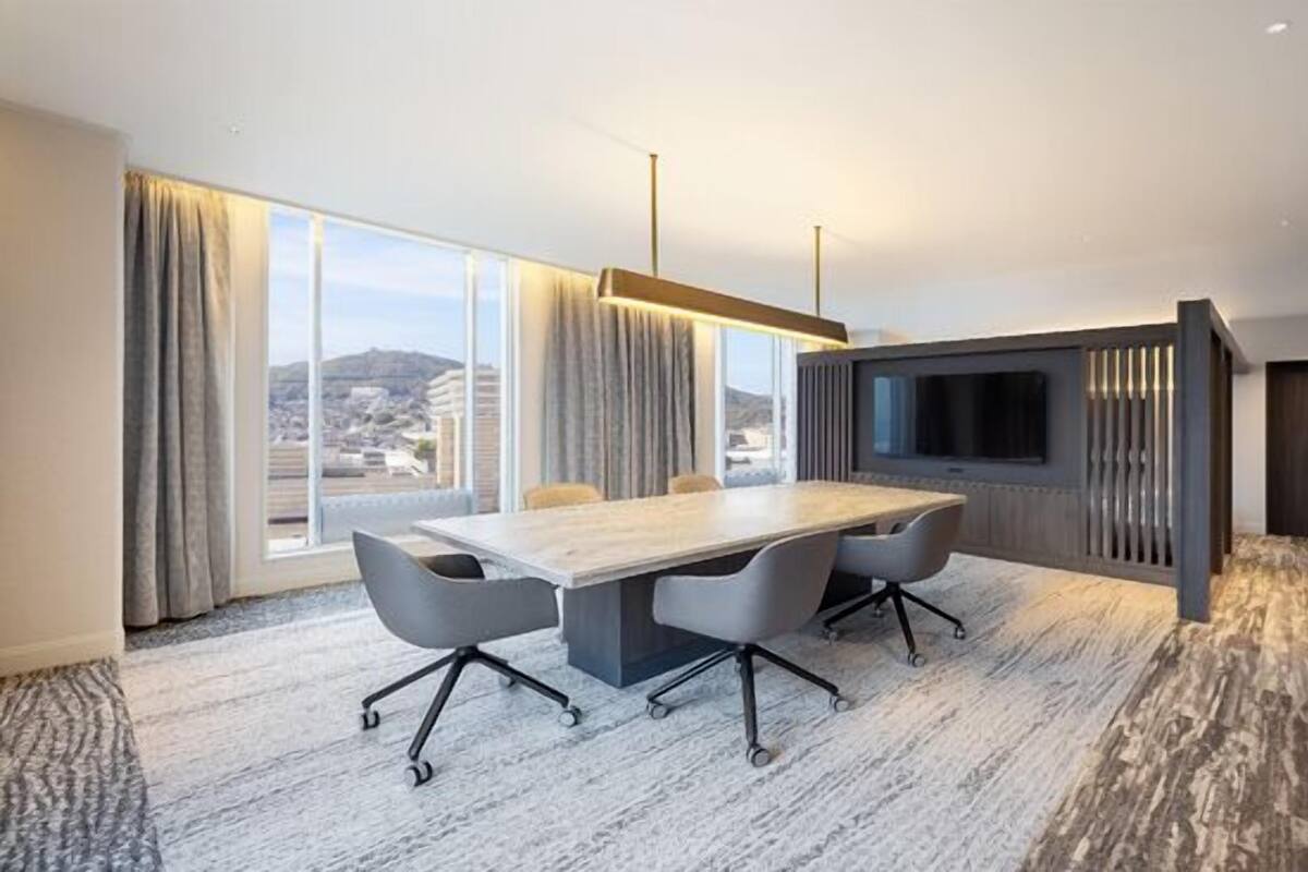 A modern hotel room with a large conference table, a flat-screen TV, and a city view.