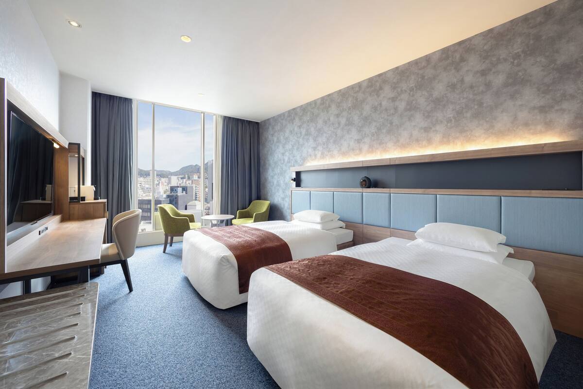 A hotel room with two beds, a desk, a chair, and a large window offering a city view.