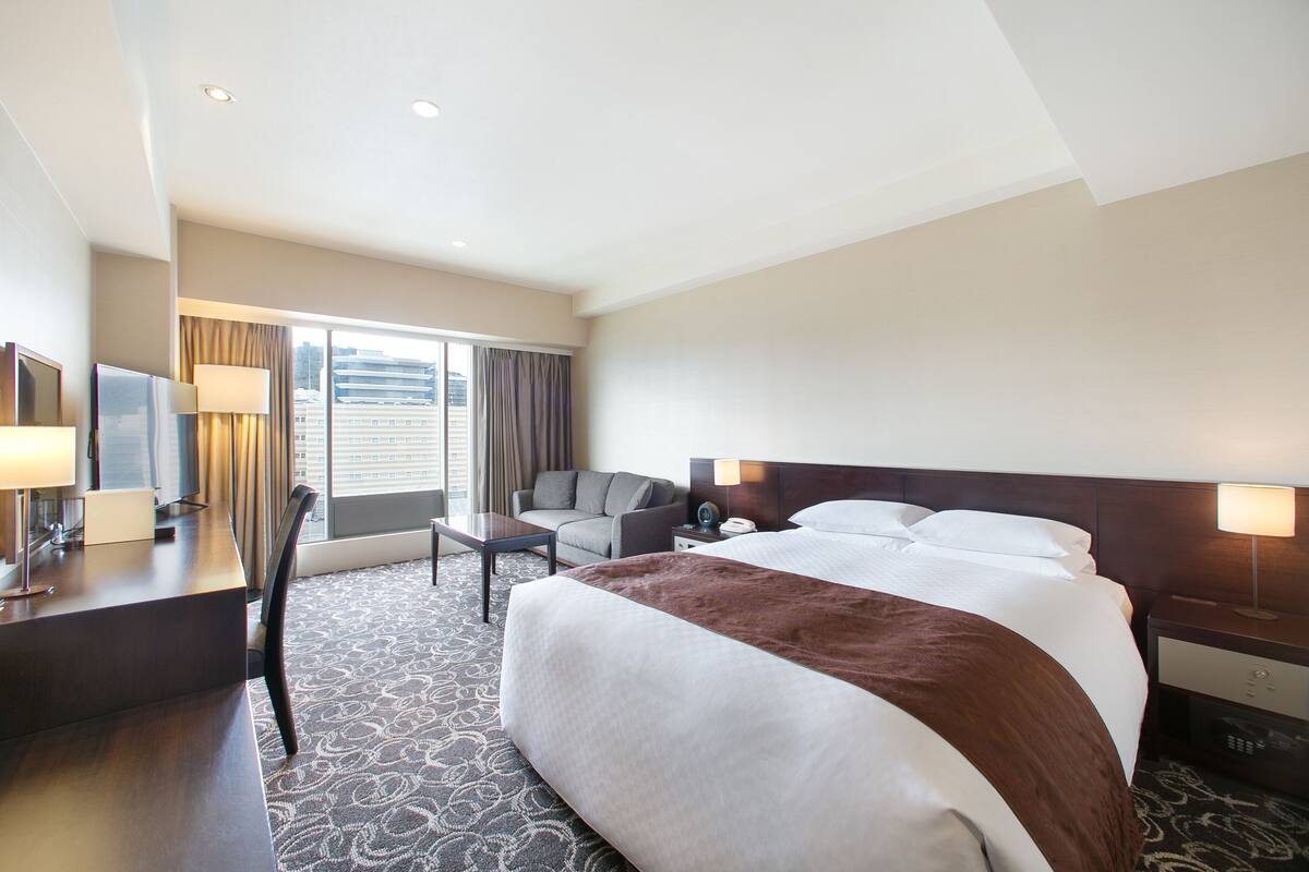 A hotel room with a large bed, a desk with a lamp, a chair, and a window with curtains.