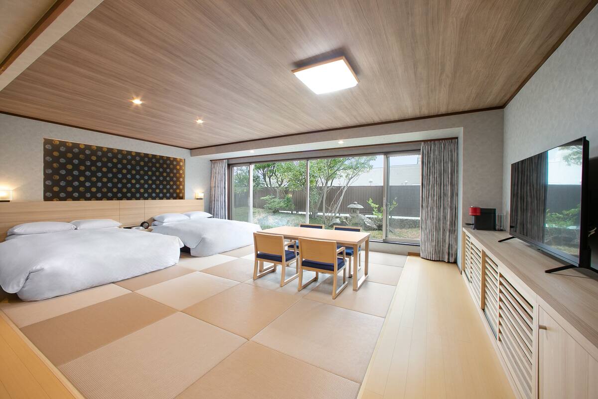 A modern hotel room with tatami flooring, a large bed, a dining table with chairs, and a TV on a wooden cabinet.