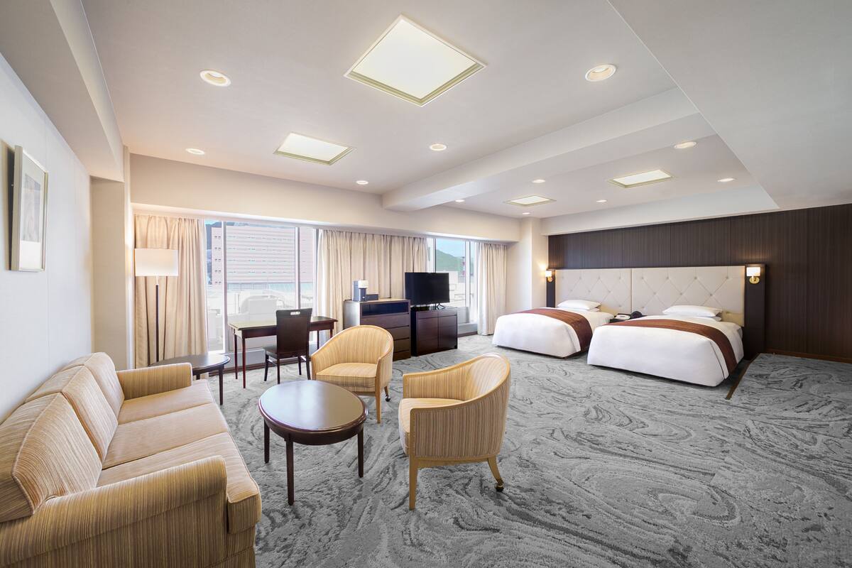 A hotel room with a sofa, two armchairs, a small table, a bed with two pillows, a TV, and a large window with curtains.