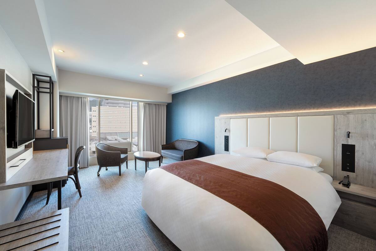 A modern hotel room with a large bed, a desk with a flat-screen TV, a chair, and a small table.