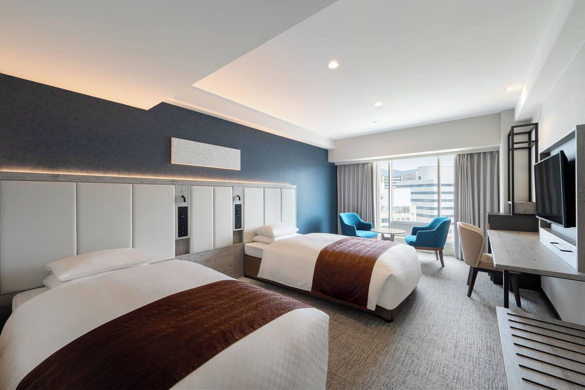 A hotel room with two beds, a desk, a chair, a TV, and a large window with curtains.