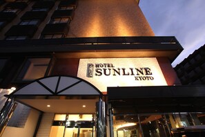 Exterior del Hotel Sunshine Kyoto con un llamativo letrero iluminado, una entrada moderna con fachada de vidrio y un cielo despejado al fondo.