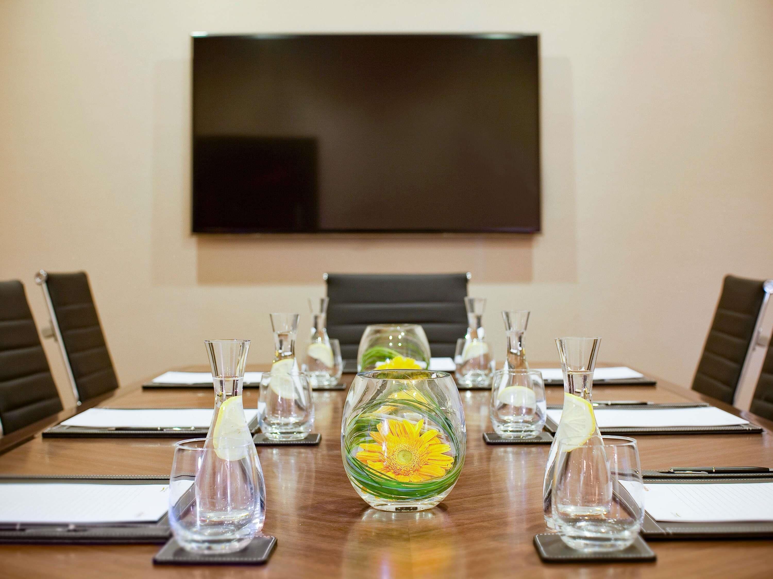 Una mesa de conferencias con vasos de agua, un centro de mesa con una flor y un televisor de pantalla plana al fondo.