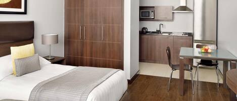 A hotel room with a bed, bedside table, lamp, wardrobe, kitchenette, and a dining area.