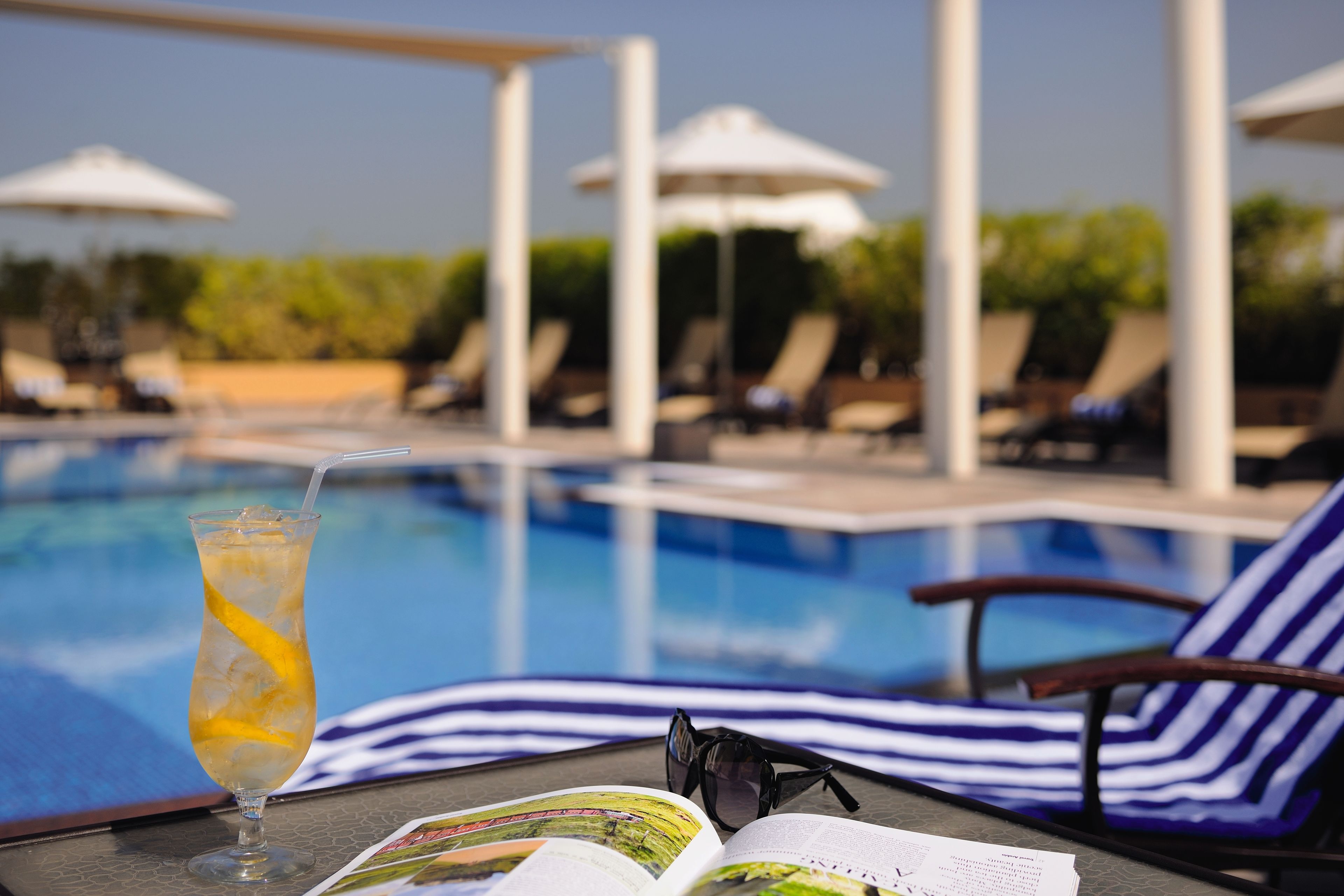 A poolside view with lounge chairs, umbrellas, and a drink on a table.