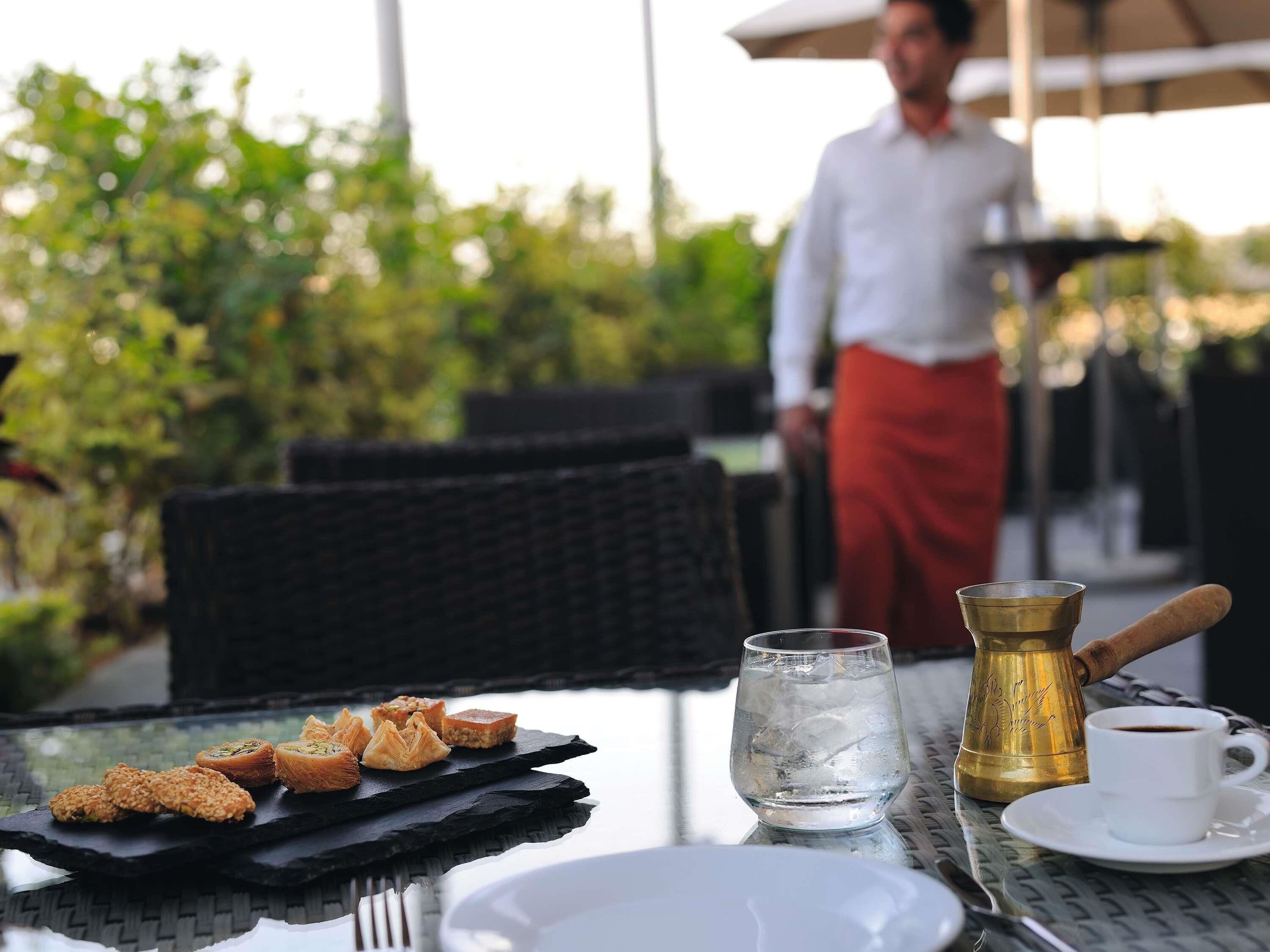 A table with a plate of pastries, a glass of water, a coffee cup, and a brass coffee pot.