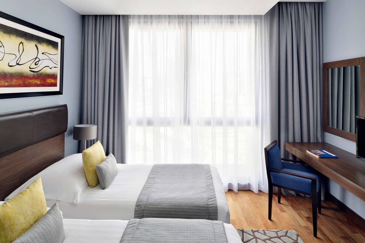 A hotel room with two beds, a desk, a chair, a TV, and a large window with curtains.