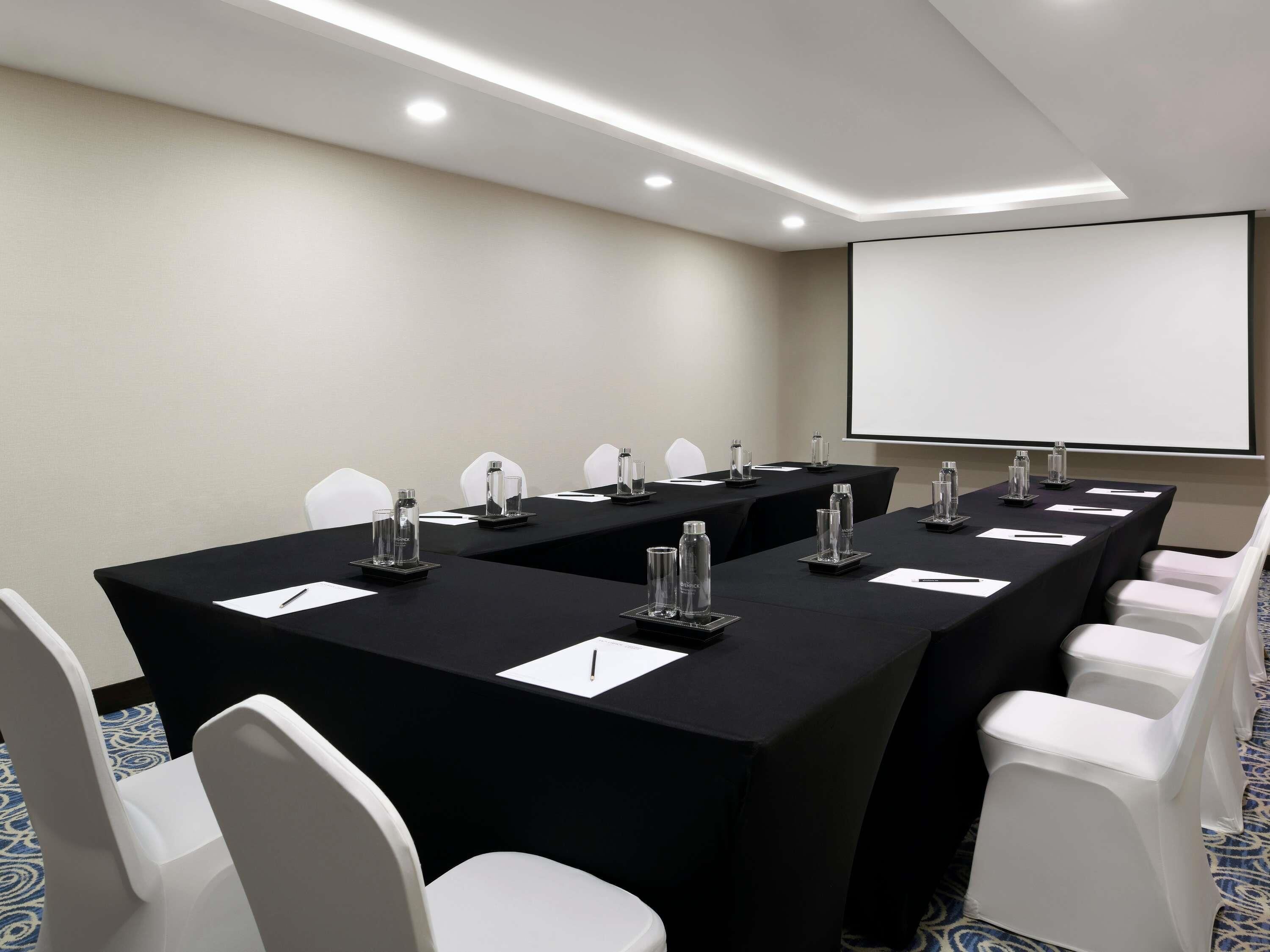 Ein Konferenzraum mit einem langen Tisch, weißen Stühlen und einer Projektionsleinwand.