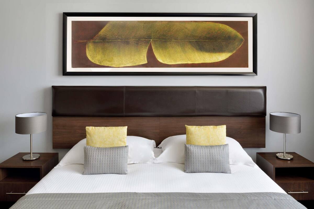 A neatly made bed with two pillows, a headboard, bedside tables with lamps, and a framed artwork above the bed.