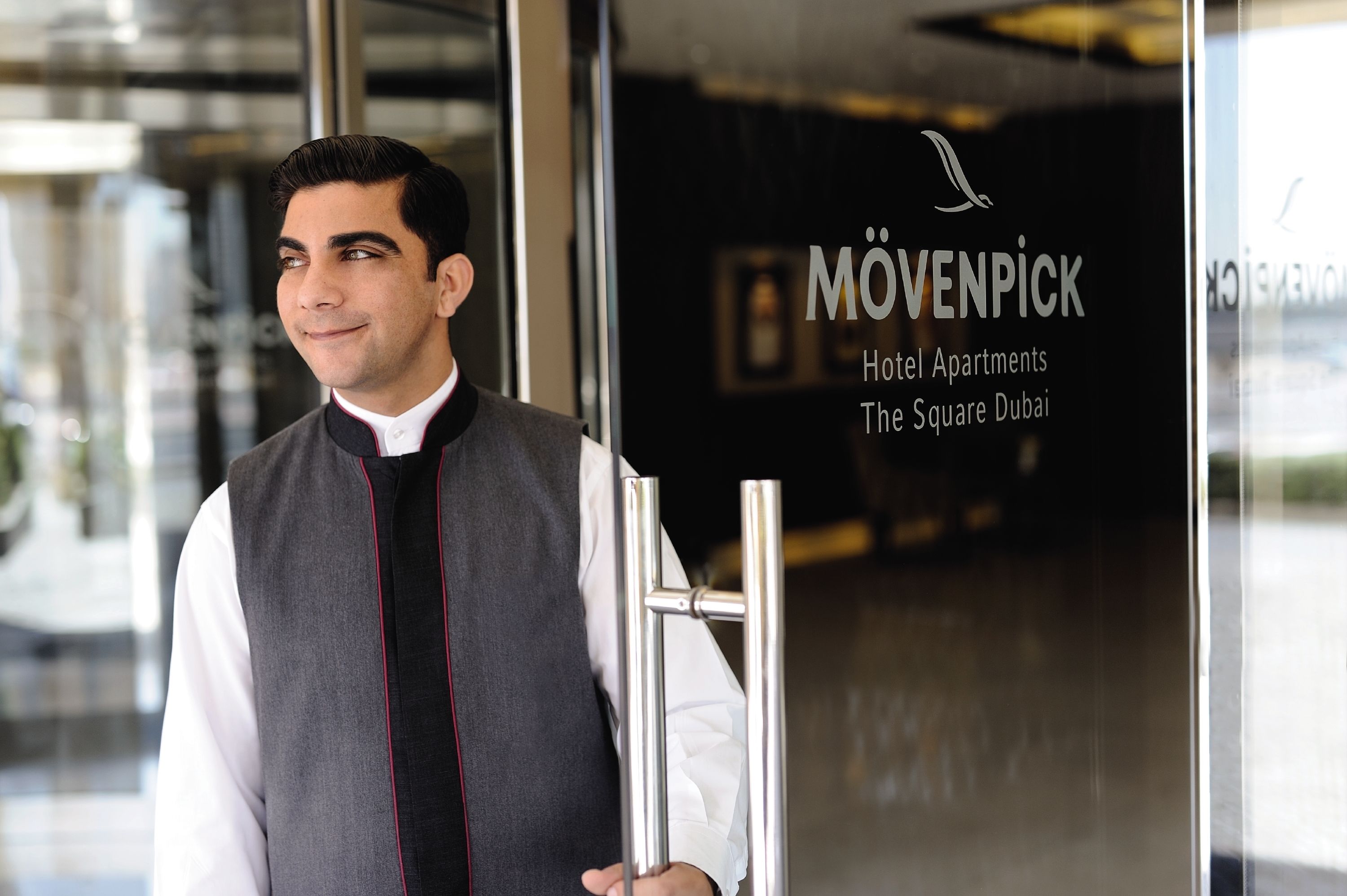 A man in a vest standing in front of a hotel entrance with a sign reading "Mövenpick Hotel Apartments The Square Dubai".