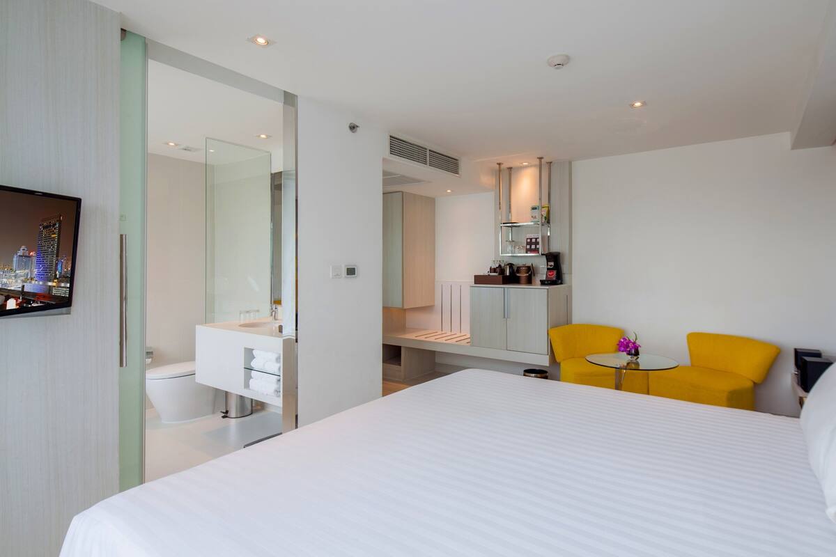 Ein modernes Hotelzimmer mit einem großen Bett, einem kleinen runden Tisch, einem gelben Sessel, einem Badezimmer mit Duschkabine aus Glas und Blick auf die Stadt auf dem Fernseher.