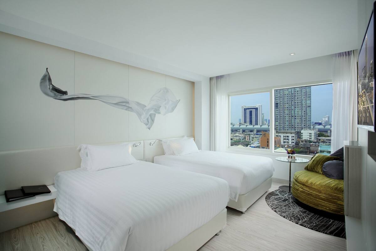 Ein Hotelzimmer mit zwei Betten, Blick auf die Stadt, einem Bild an der Wand und einem kleinen Tisch mit einer Vase.