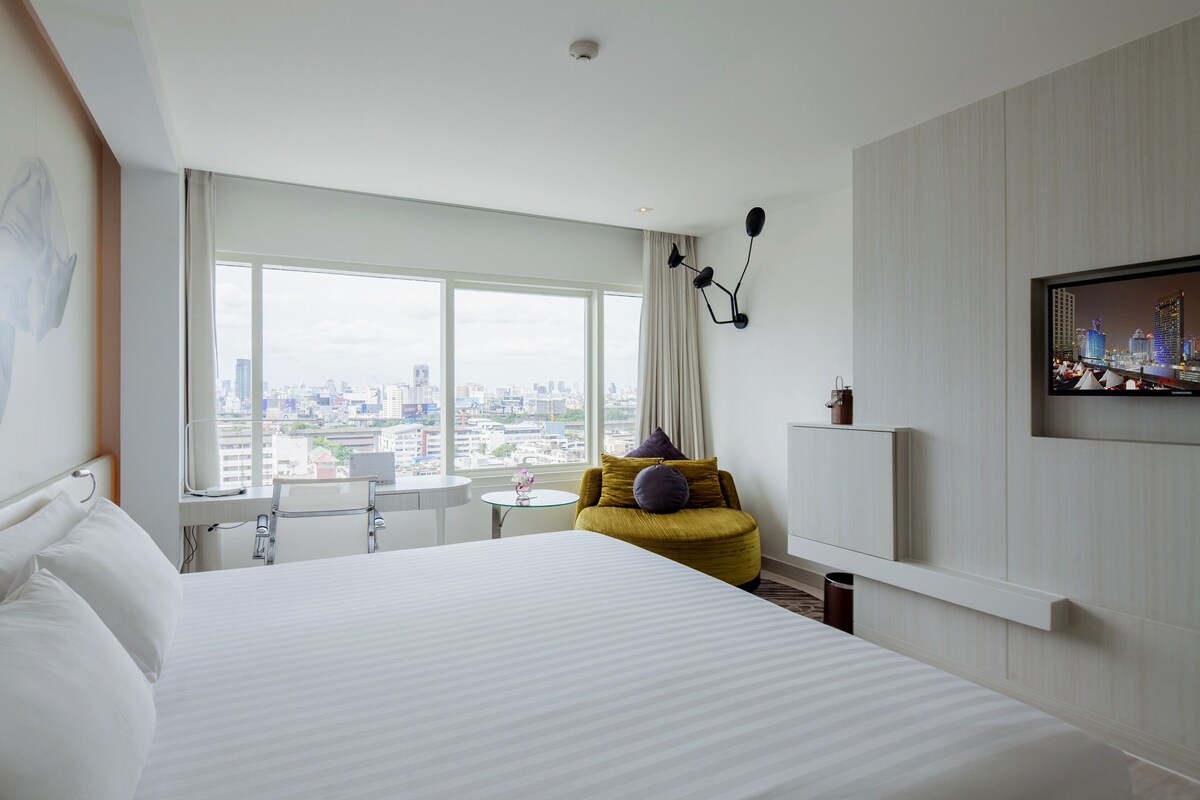 Habitación de hotel con una cama grande, un escritorio, una silla, una pequeña mesa con una flor, un sofá con cojines, un televisor en la pared y vistas a la ciudad.