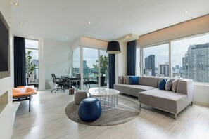Un salon moderne avec un canapé, une table basse ronde et un fauteuil. On y trouve également une table à manger avec des chaises, ainsi qu’une grande fenêtre donnant sur la ville.