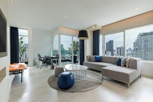 Uma sala de estar moderna com um sofá, uma mesa de centro redonda e uma poltrona. Há uma mesa de jantar com cadeiras e uma janela grande com vista para a cidade.
