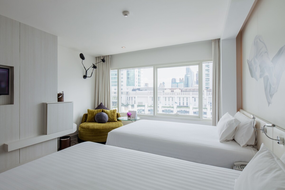 Habitación de hotel con dos camas, televisión, escritorio con lámpara y ventana grande con vista a la ciudad.