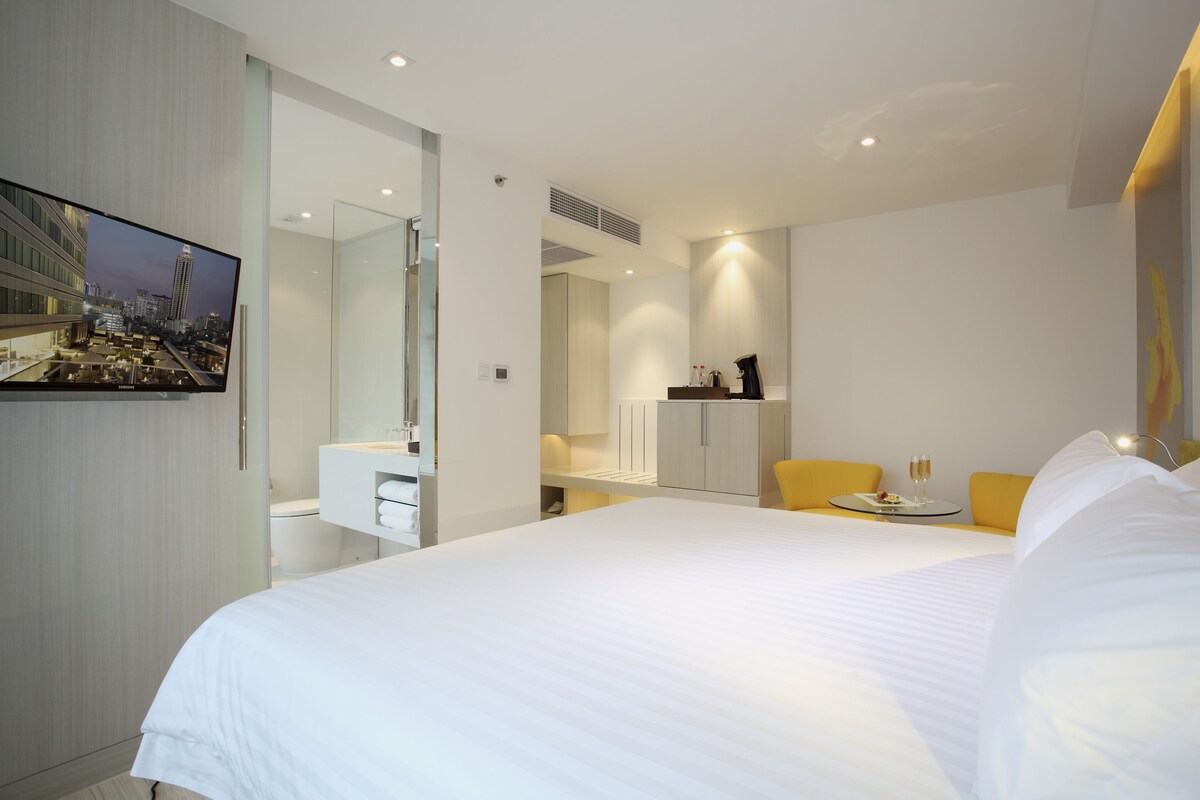 Una habitación de hotel moderna con una cama grande, un televisor de pantalla plana en la pared, un baño con espejo y una mesita con un vaso de agua.