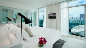 Une chambre d’hôtel moderne dotée d’un grand lit, offrant une vue sur la ville, d’un téléviseur fixé au mur et d’un espace salon circulaire.