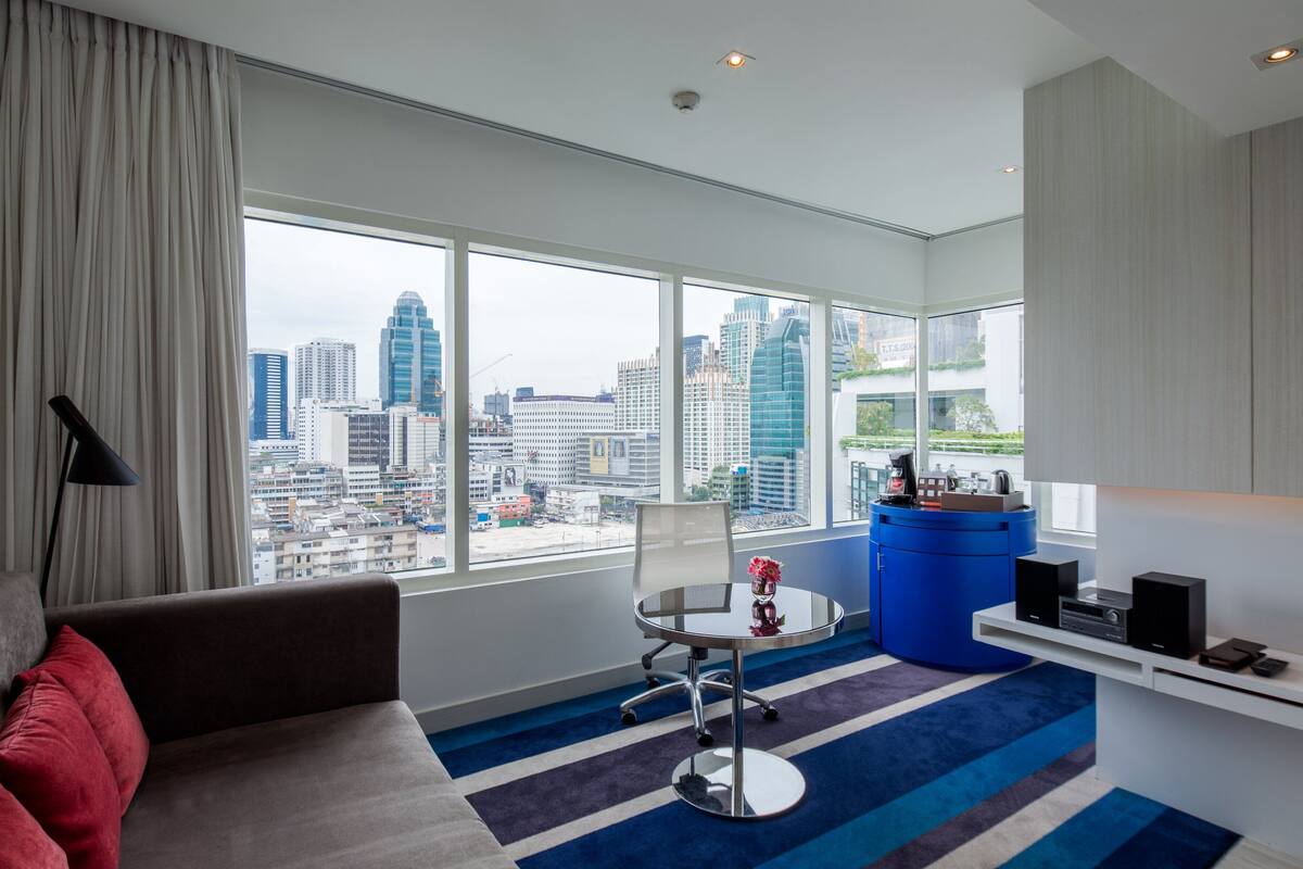 Ein modernes Hotelzimmer mit großem Fenster und Blick auf die Stadt, einem runden Tisch, einem blauen Schrank und einer Couch mit roten Kissen.