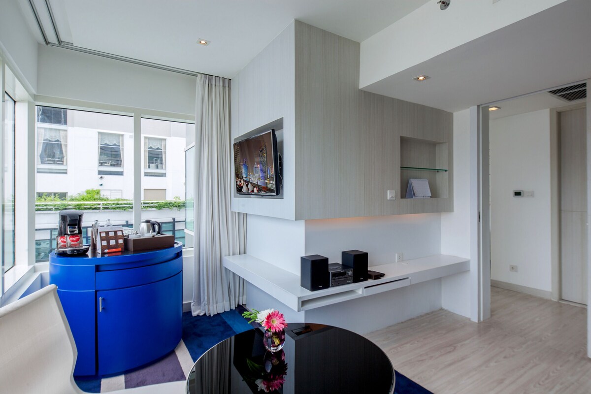 Una habitación de hotel moderna con un ventanal grande, un minibár azul, un televisor de pantalla plana montado en la pared y un escritorio blanco.