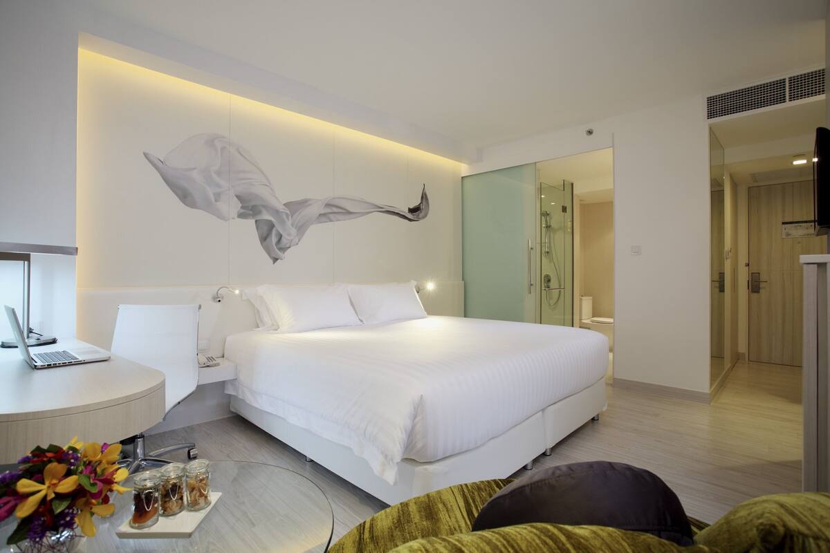 Ein modernes Hotelzimmer mit einem großen Bett, einem Schreibtisch mit einem Laptop, einer Duschkabine aus Glas und einer minimalistischen Einrichtung.