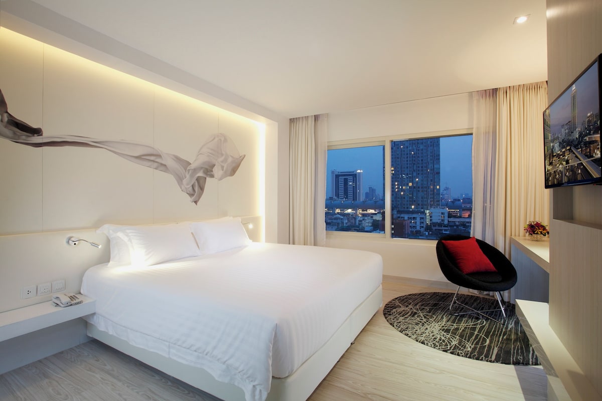 A modern hotel room with a large bed, a city view, a red chair, and a wall-mounted artwork.