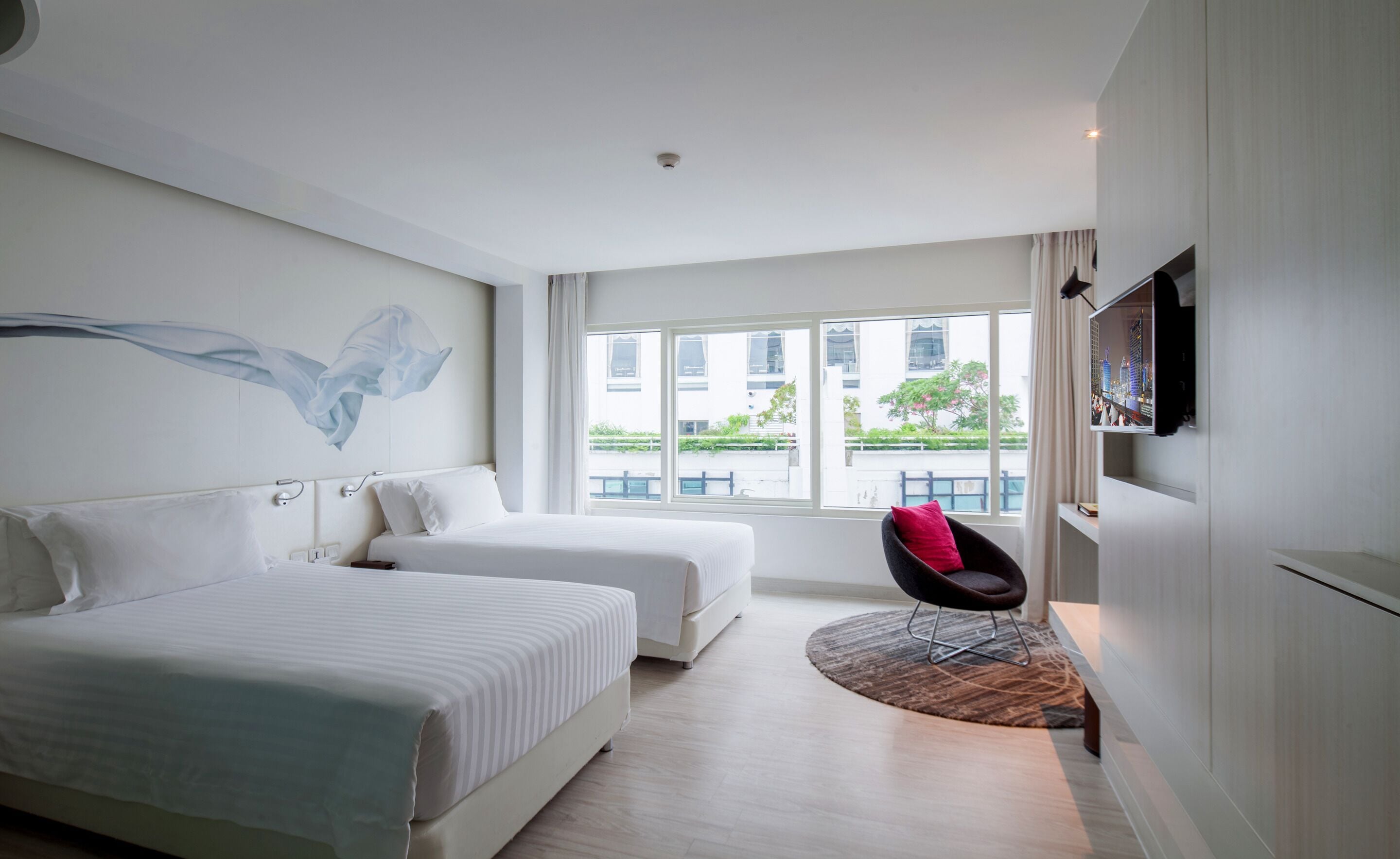 A modern hotel room with two beds, a large window with a view of buildings, a mounted TV, and a unique chair with a red cushion.