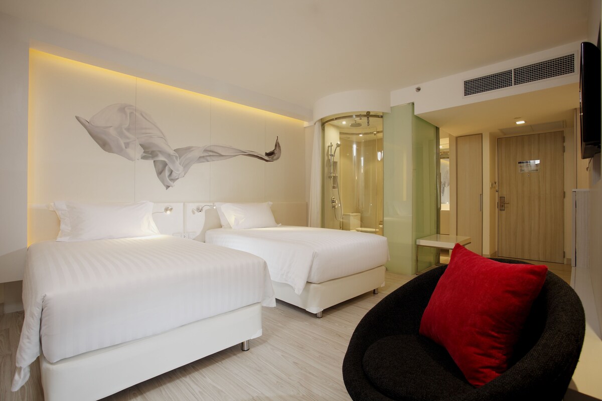 Habitación de hotel con dos camas, zona de ducha, televisor y almohada roja.